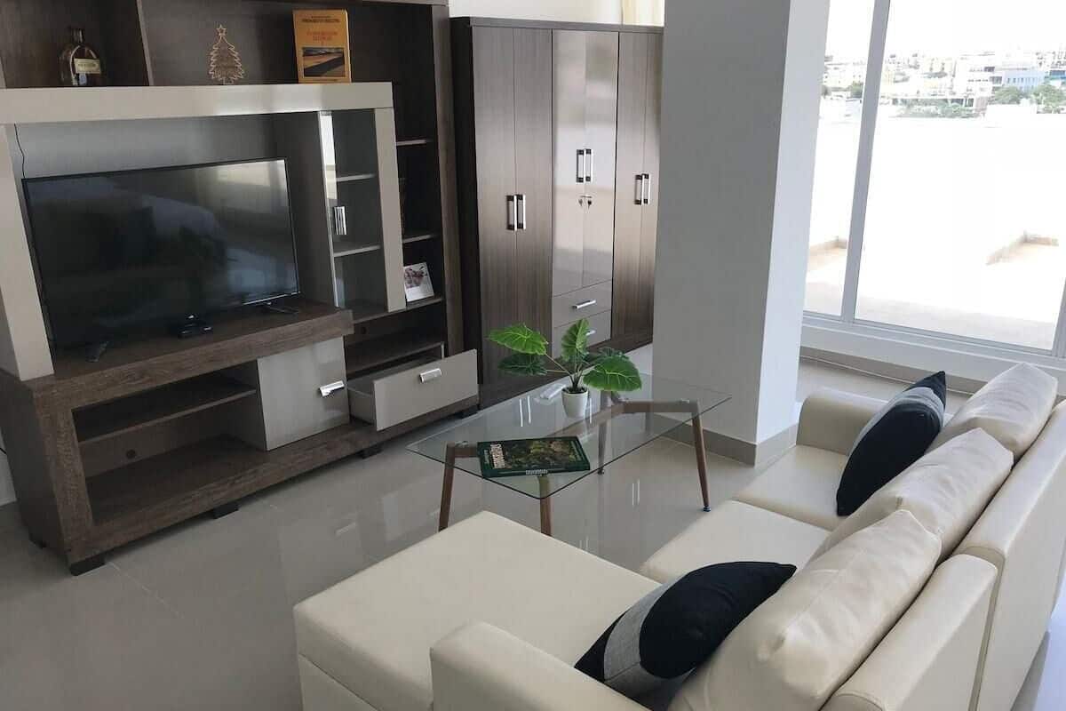 A modern living room with a flat-screen TV, a glass coffee table, a sofa with cushions, and a cabinet with drawers.