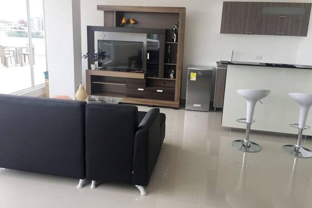 A modern living room with a black sofa, a TV, and a kitchen area with a fridge and bar stools.