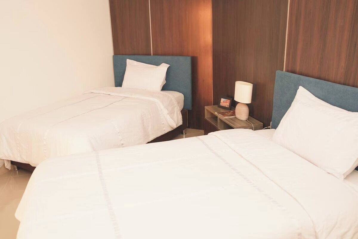 A hotel room with two beds, each with white bedding and blue headboards. There is a nightstand with a lamp and a small decorative item.