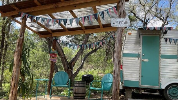 Una pérgola de madera con un letrero que dice 'Welcome', dos sillas verdes, un barril y una caravana vintage.