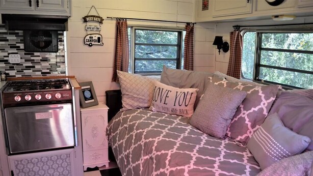 A compact RV interior with a kitchenette, a bed with patterned bedding, and a window with a view of trees. - The Blue Moon on Hamilton Creek (Burnet County)
