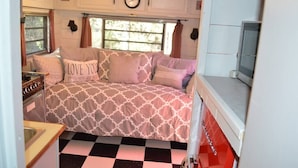 A cozy bedroom with a checkered floor, a bed with patterned bedding, a clock on the wall, and a window with curtains. - The Blue Moon on Hamilton Creek (Burnet County)