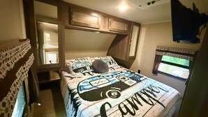 A bedroom with a bed, a window, a ceiling fan, and a TV mounted on the wall.