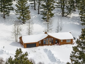 Una cabaña de troncos con techo nevado, rodeada de árboles, y un cielo despejado.