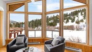 A cozy room with large windows, two leather chairs, a small table, and a view of a snowy landscape. - Cabin near Bridger Bowl with Hot Tub!  (Bozeman)