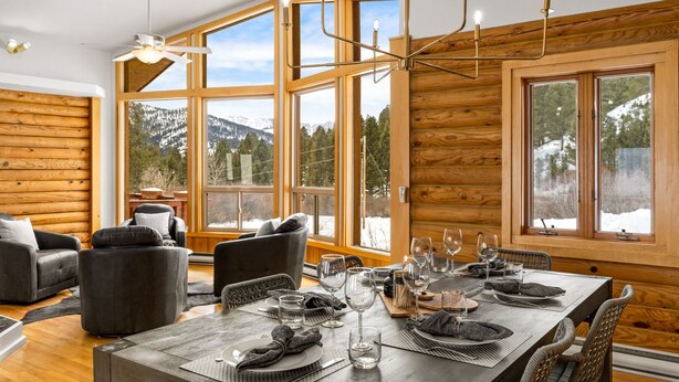 A cozy log cabin interior with a dining area set for four, large windows offering mountain views, and a ceiling fan. - Cabin near Bridger Bowl with Hot Tub!  (Bozeman)