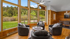 A spacious living room with large windows, a ceiling fan, and a view of a forested landscape. - Cabin near Bridger Bowl with Hot Tub!  (Bozeman)