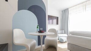 A modern living room with a round table, two white chairs, a sofa, and a large abstract wall mural. - zzzpace Smart-Hotel Bielefeld (Bielefeld)