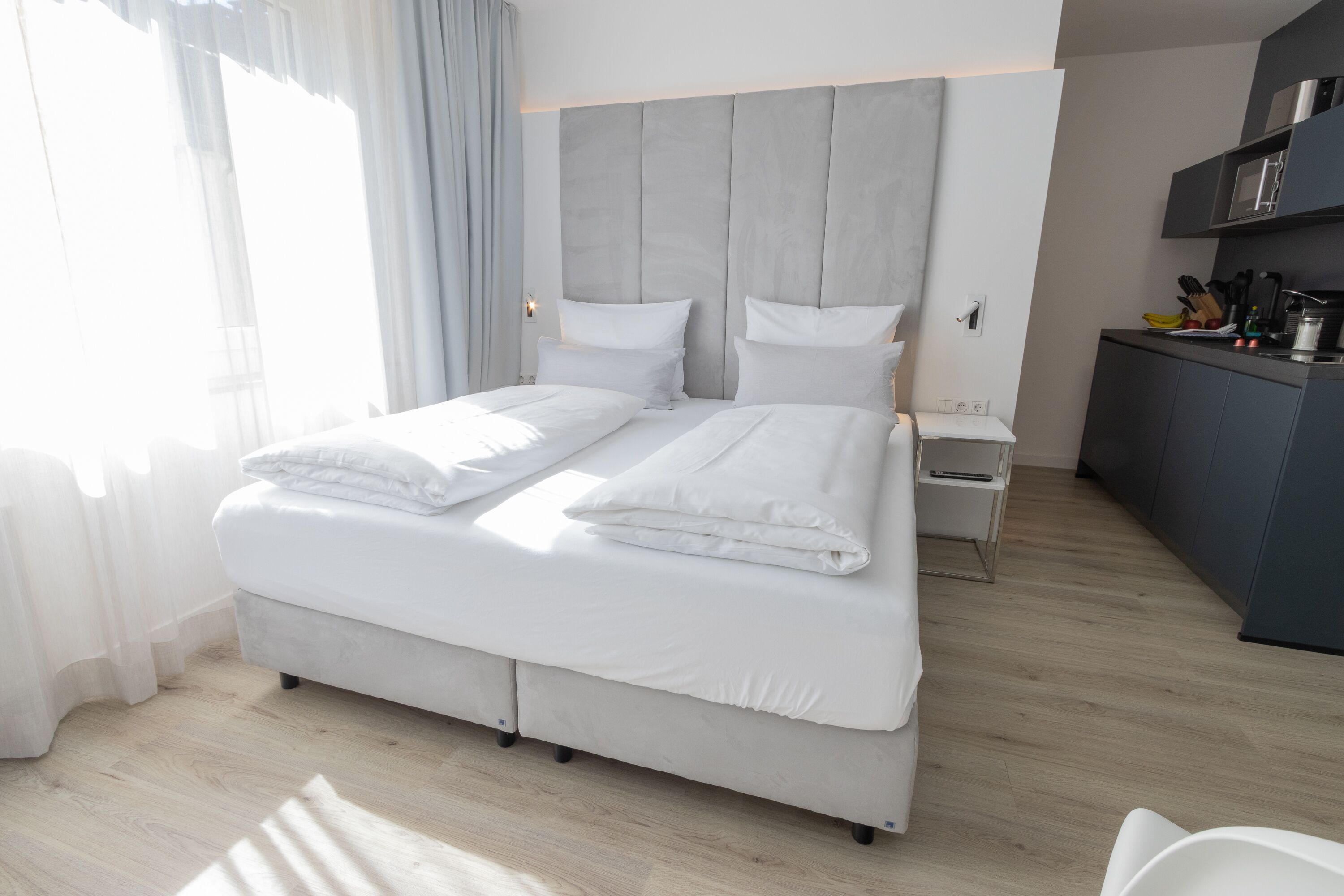 A modern hotel room with a large bed, a bedside table, a built-in desk with a chair, and a view of the city.