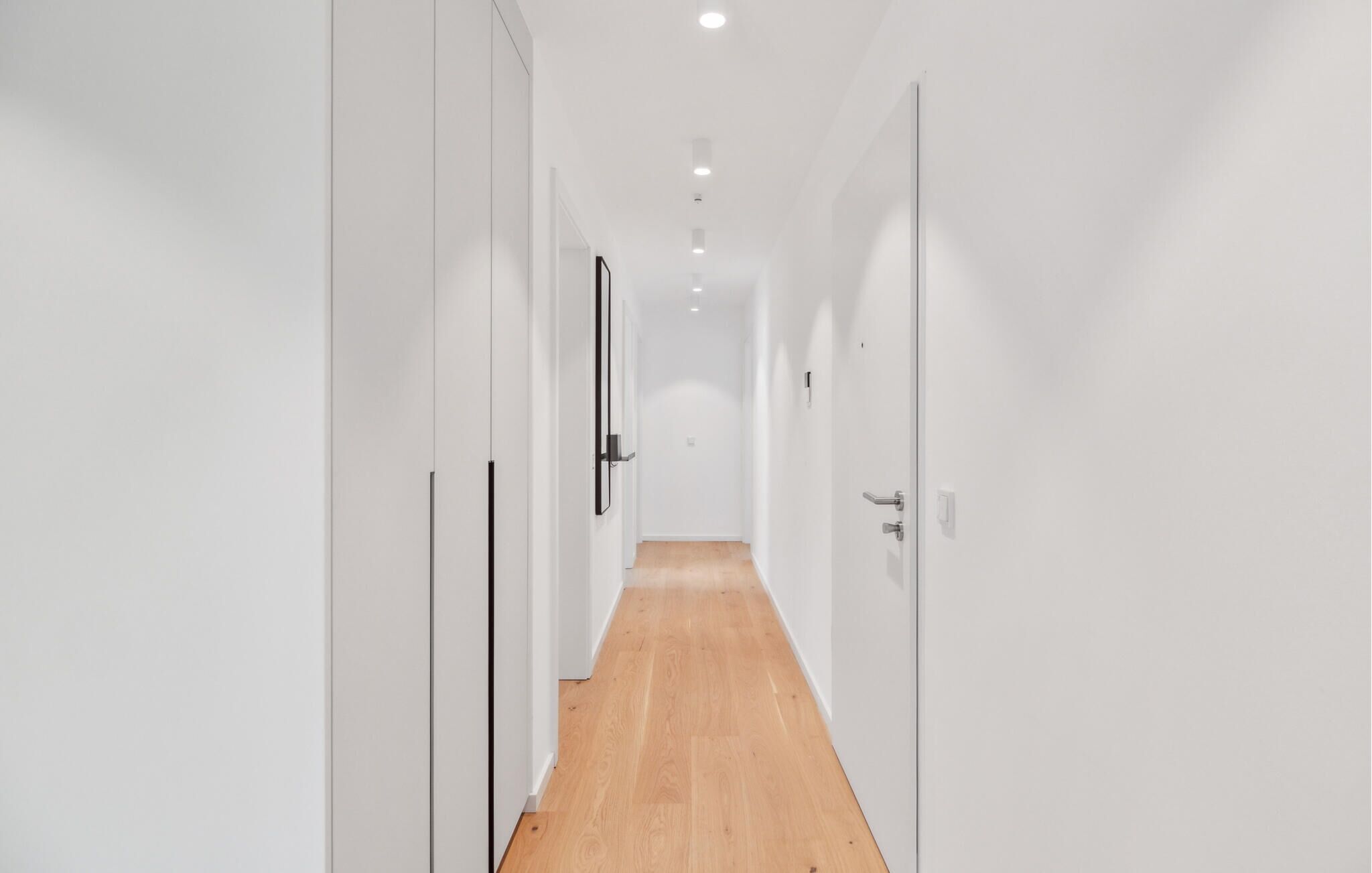 A narrow, well-lit hallway with white walls and wooden flooring, leading to a door at the end.