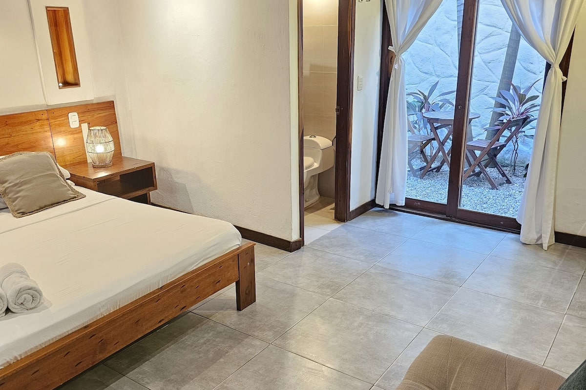 A bedroom with a wooden bed, a nightstand, a bedside lamp, and a view of the bathroom through a glass door.