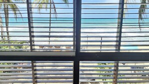 Vista a través de persianas de una playa con palmeras y un cielo azul despejado.