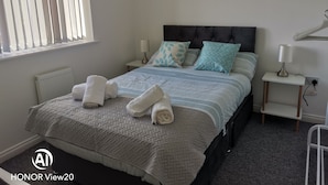 A neatly made bed with a quilted bedspread, two pillows, and rolled towels on a headboard. There is a bedside table with a lamp, a wall-mounted painting, and a radiator. - Comfortable 3 BR with parking-WS2 for contractors, professionals and families
 (Walsall)