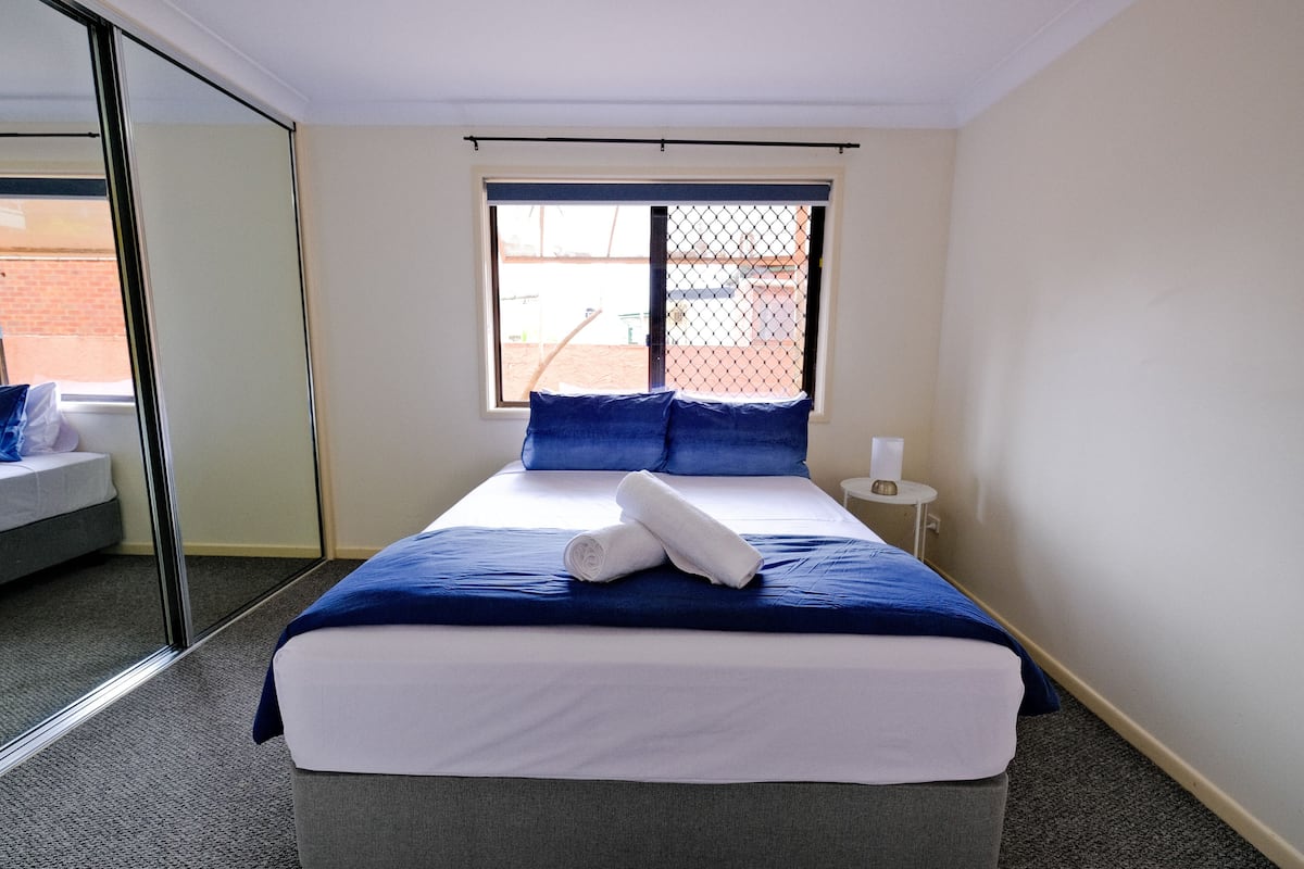 A neatly made bed with a blue and white duvet, a rolled white towel, and a small bedside table with a lamp.
