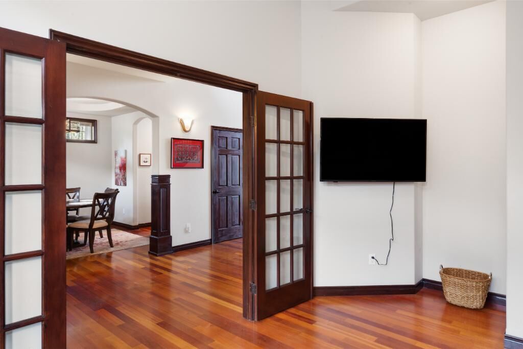 Una habitación con suelo de madera, una puerta de madera, un televisor de pantalla plana montado en la pared y un comedor visible a través de un puerta abierta.