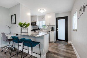Una cocina moderna con armarios blancos, encimera de granito y microondas integrado. Hay dos taburetes de bar color turquesa y una silla de oficina gris.
