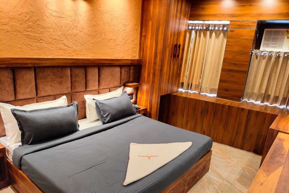 A hotel room with a wooden headboard, a bed with dark bedding, a bedside table, and a window with curtains.