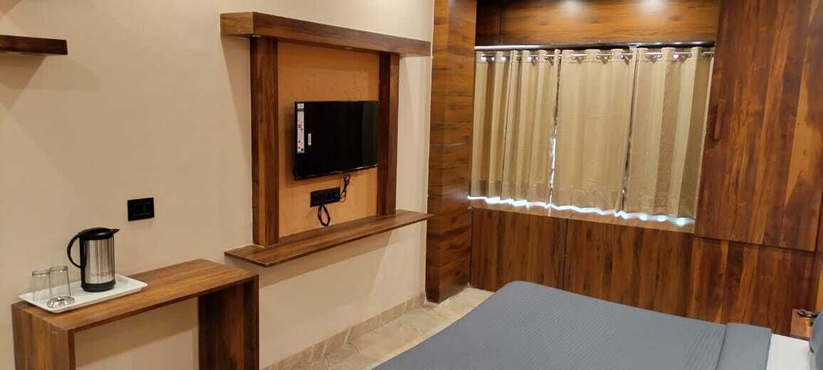 A small room with a wooden wall, a television mounted on a wooden shelf, a window with curtains, a bed, and a small table with a kettle and glasses.