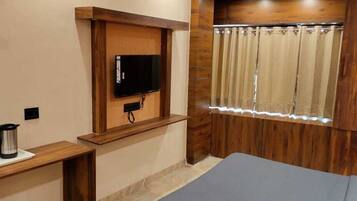 A small room with a wooden wall, a television mounted on a wooden shelf, a window with curtains, a bed, and a small table with a kettle and glasses.