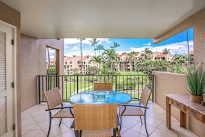 A balcony with a glass table and wicker chairs, overlooking a landscaped area with palm trees and buildings. - Affordable, Luxury Resort condo w/ AC @ Kam III Beach  (Kihei)