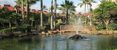 A serene pond with a fountain, surrounded by palm trees and a wooden bridge.