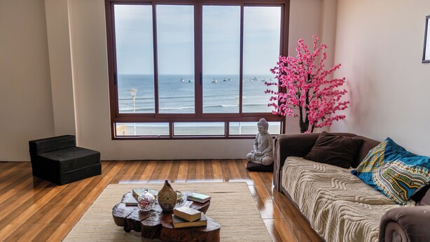 A room with a large window overlooking the sea, a sofa, a small table with books and decorative items, and a statue. - Hotel Sueños de Chicama (Rázuri)