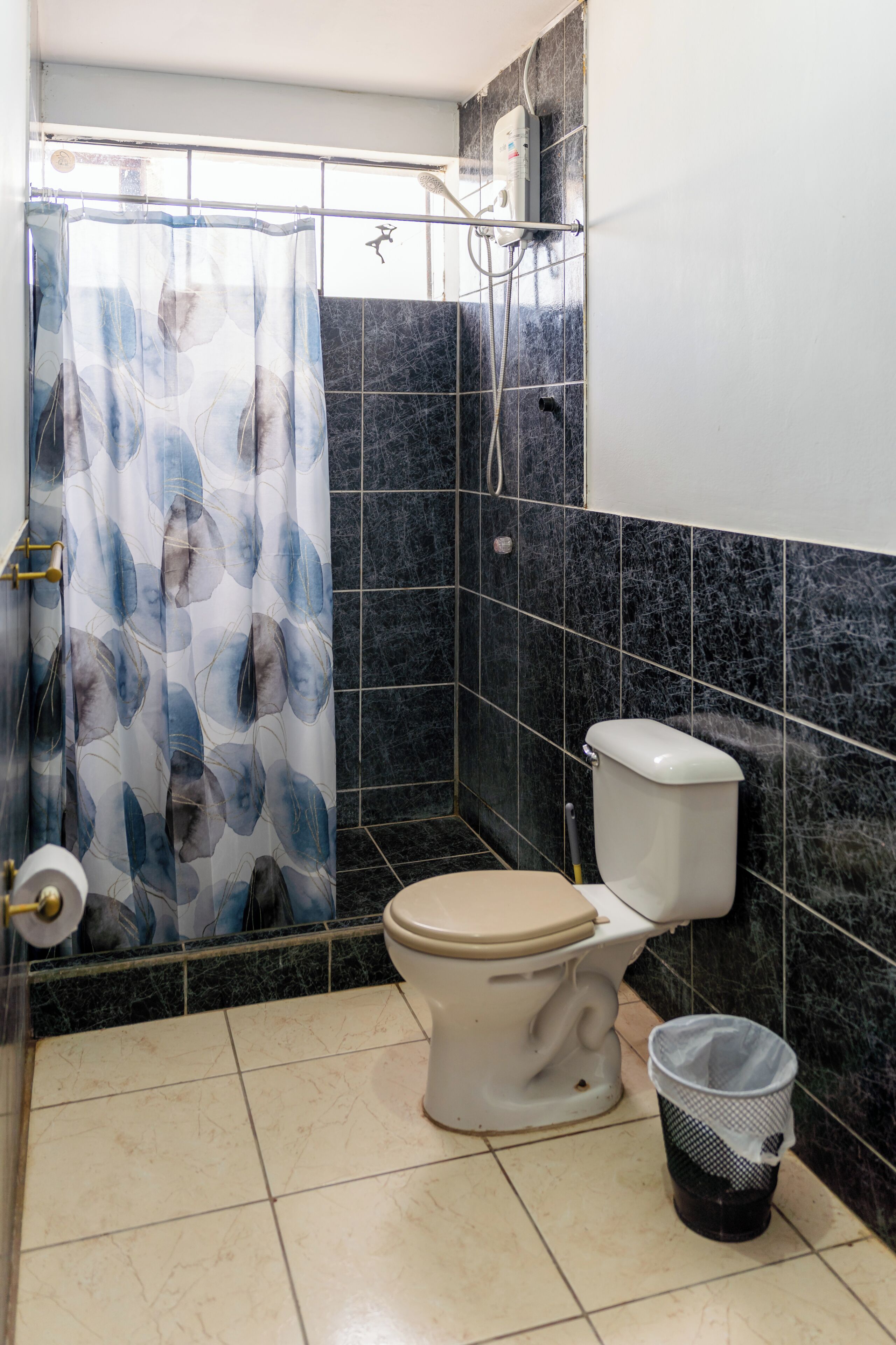 Une salle de bain avec des toilettes, un rideau de douche, une poubelle et un carrelage au sol.