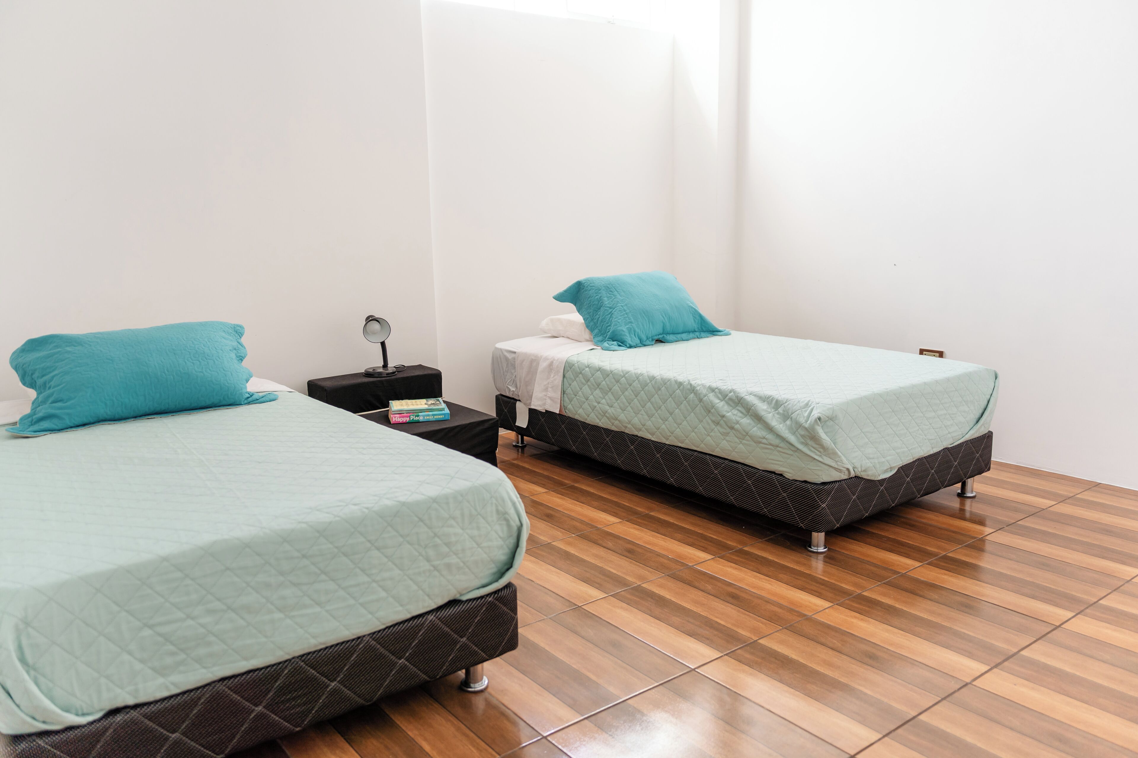 Deux lits simples avec une literie de couleur turquoise, une table de chevet avec une lampe et un plancher en bois.