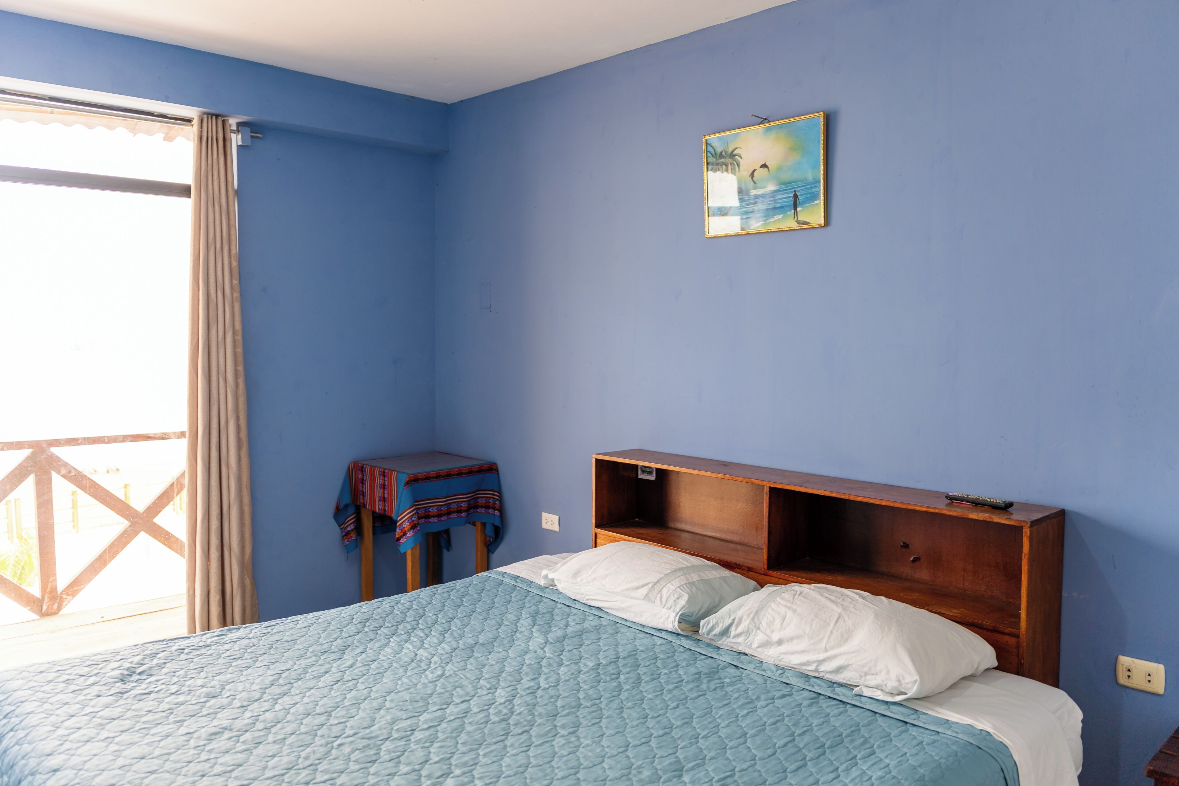 Quarto com cama, cabeceira de madeira, mesa pequena com tecido, pintura na parede e janela com cortinas.