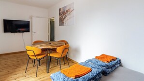 Une pièce comprenant une table à manger en bois, des chaises orange, un canapé recouvert de couvertures bleues et oranges, un téléviseur à écran plat fixé au mur et une porte donnant sur une autre pièce.