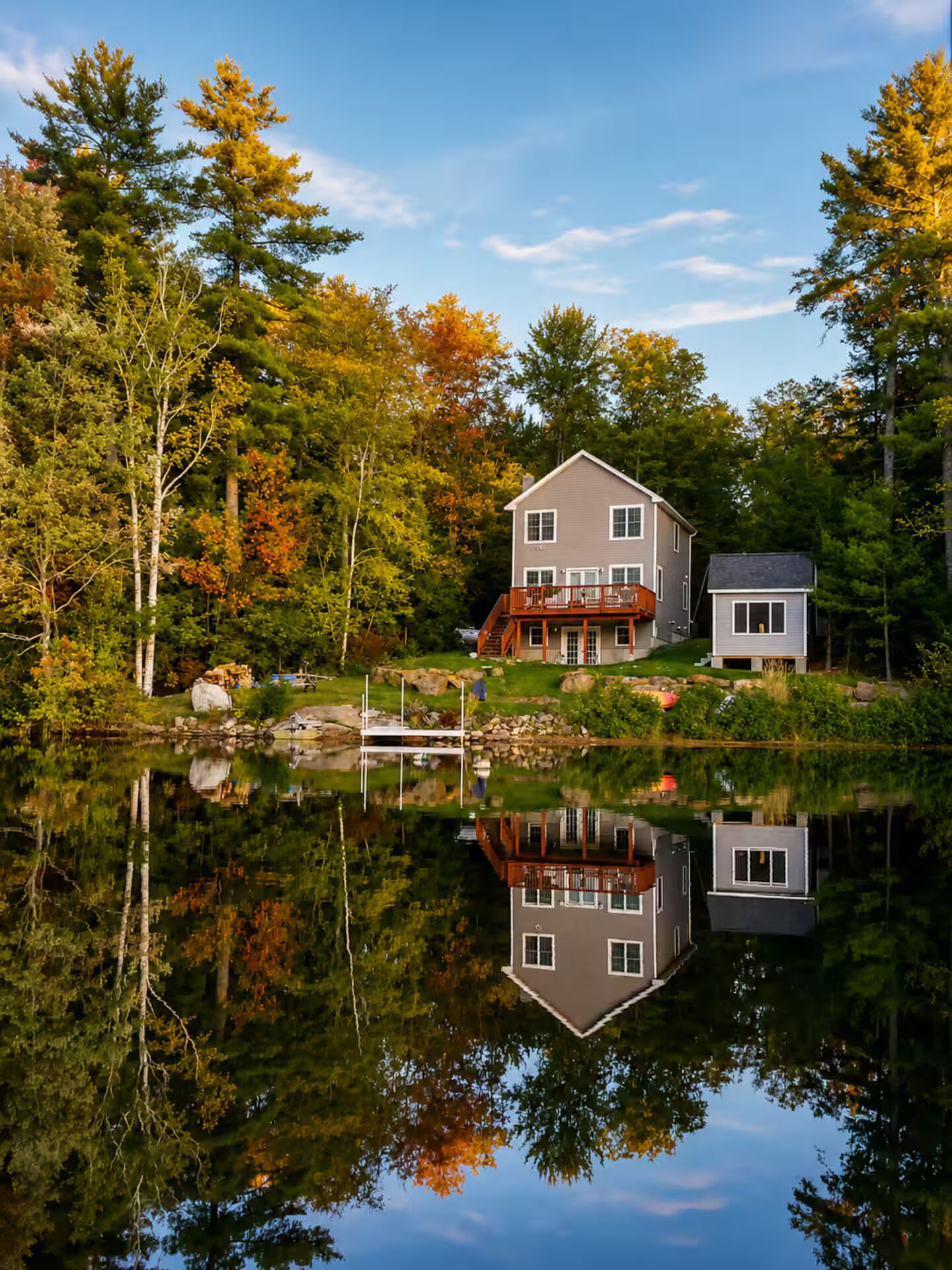 Une maison à deux étages avec un quai, entourée d’arbres et bordant un lac paisible.