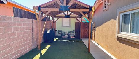 Uma área de pátio coberta com pérgola de madeira, parede de tijolos e uma janela à direita.