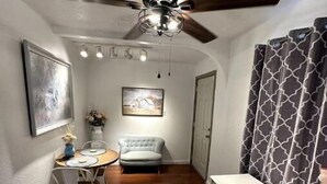 Une pièce étroite avec un ventilateur de plafond, une table à manger avec des chaises, un canapé, un bureau avec une chaise et un rideau à motifs.
