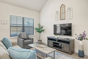 Un salon avec un canapé gris, une table basse, un téléviseur posé sur un meuble et une plante.