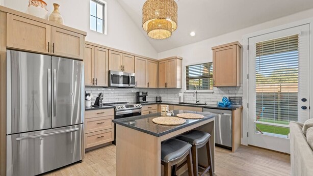 A modern kitchen with stainless steel appliances, a central island with stools, and a large window with blinds. - *New Romantic Getaway|Hot Tub (Fredericksburg)