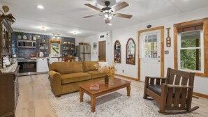 A spacious living room with a sofa, coffee table, and rocking chair. A kitchen with modern appliances and a dining area with a table and chairs are visible.