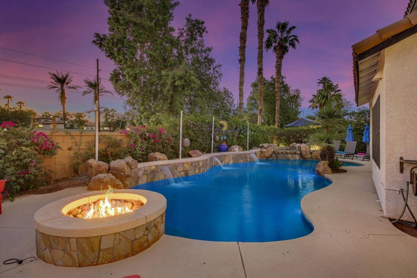 A swimming pool with a fire pit, surrounded by palm trees and a stone wall.