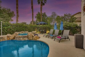 A pool area with lounge chairs and umbrellas, surrounded by greenery and palm trees. - Desert Pearl: Pool, Spa, Pool Table, Foosball, EV! (Indio)