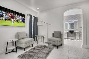 A modern living room with a sofa, armchair, and a small table. There is a large TV mounted on the wall displaying a sports event. - Desert Pearl: Pool, Spa, Pool Table, Foosball, EV! (Indio)