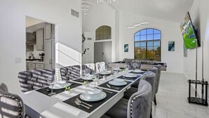 A dining area with a long table set for six, a kitchen in the background, and a large window with a view. - Desert Pearl: Pool, Spa, Pool Table, Foosball, EV! (Indio)