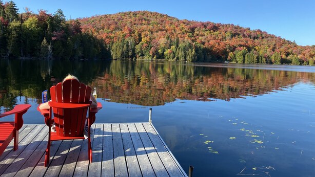 A wooden dock with a red chair and table, overlooking a calm lake with autumn foliage and a clear sky.