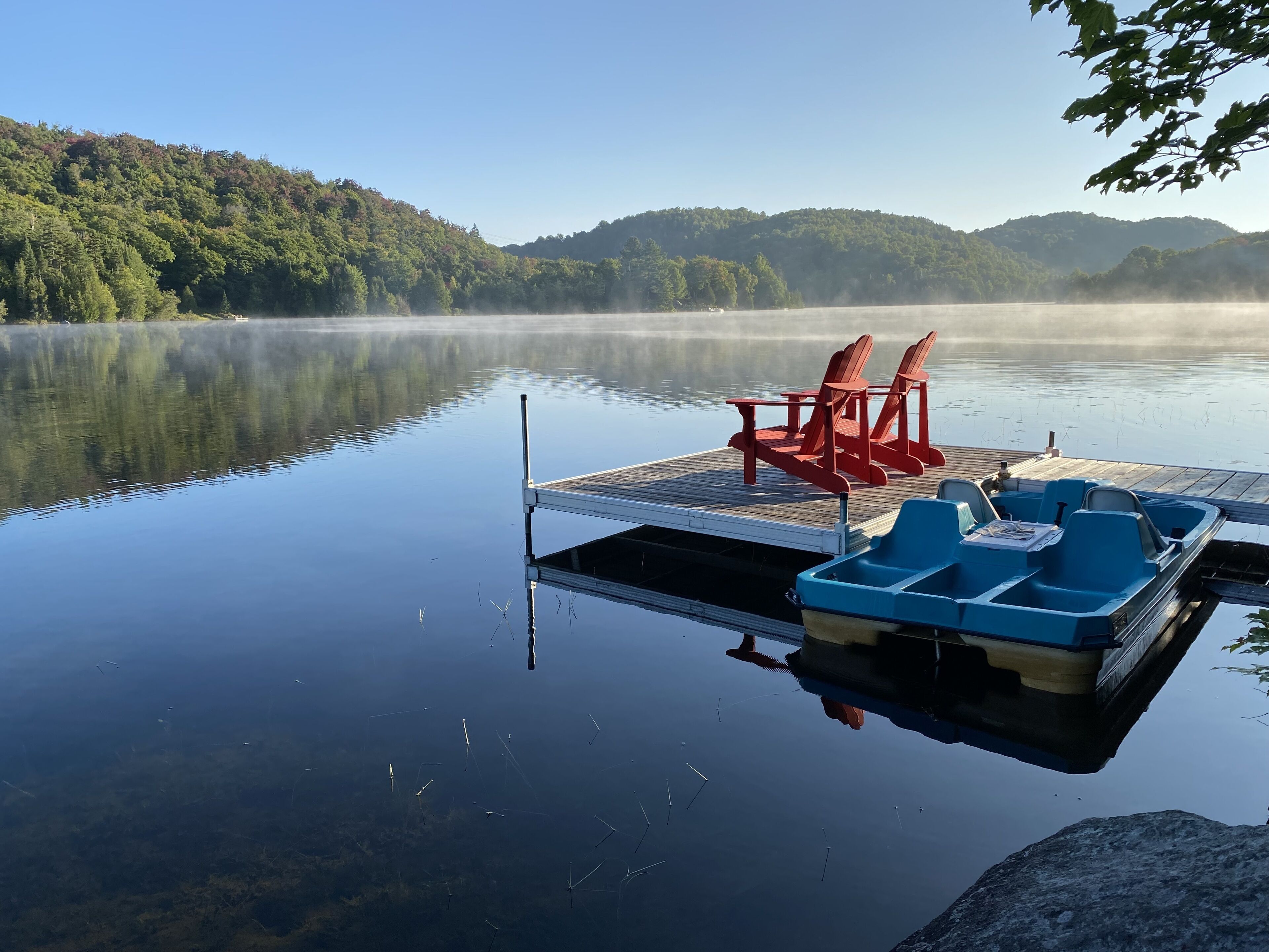 A calm lake with a wooden dock, red and blue paddle boats, and chairs.
