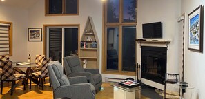 A living room with a fireplace, a television mounted above it, and a dining area with a table and chairs.