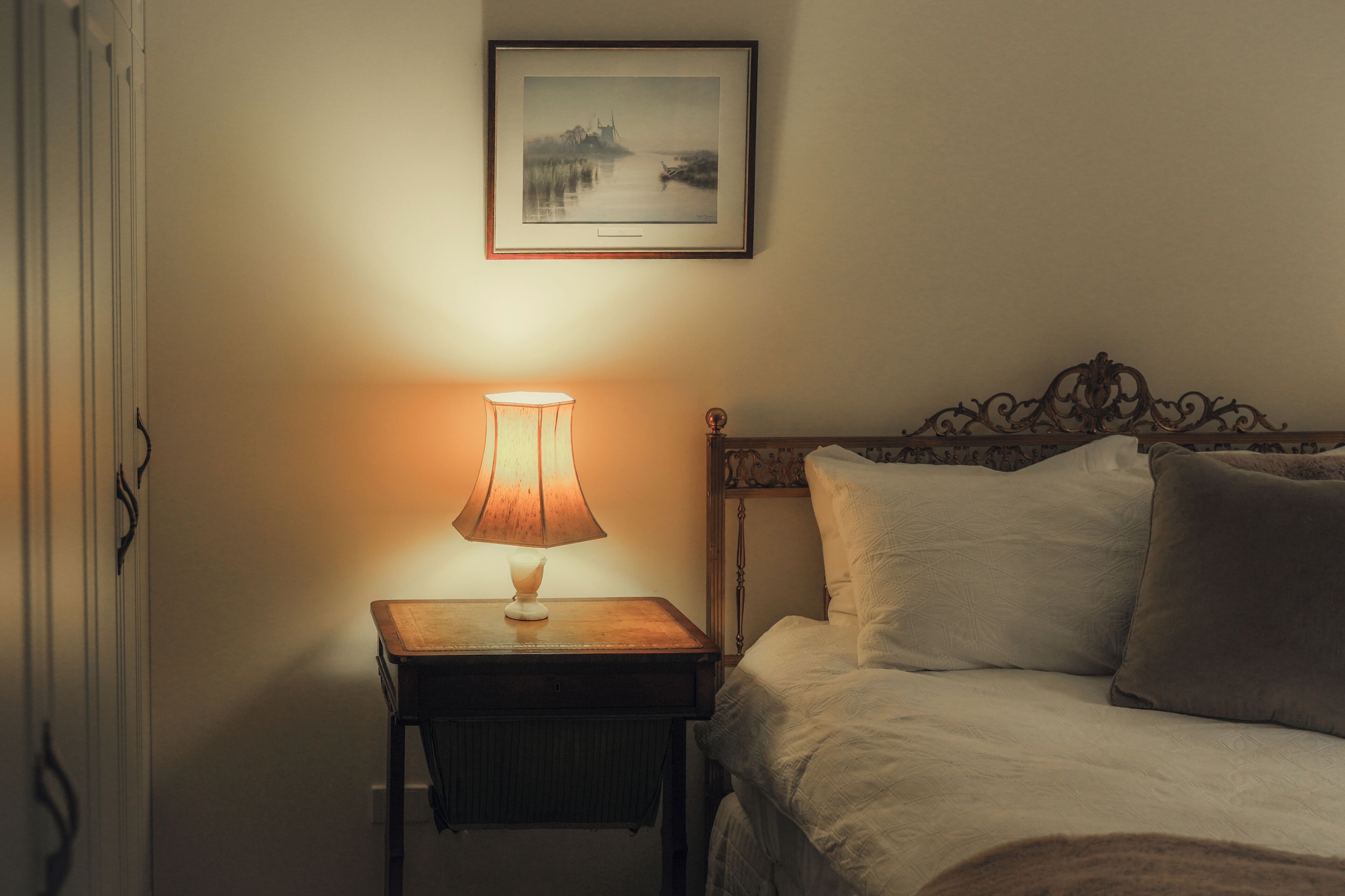 A neatly made bed with white linens and two pillows, a bedside table with a lamp, and a framed picture on the wall.
