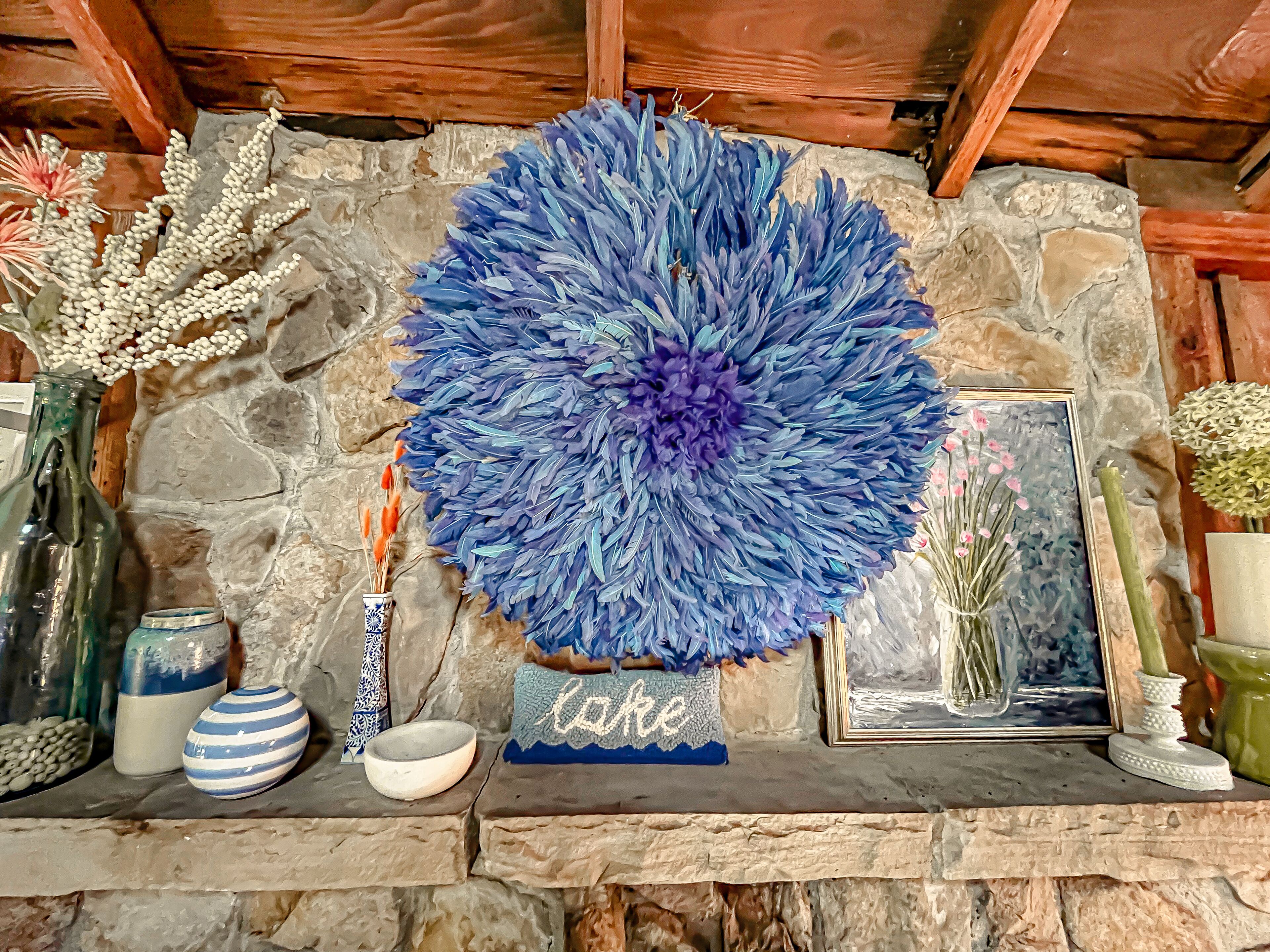 A stone fireplace with a decorative blue feather arrangement, a small sign reading "lake," and various vases and bowls on the mantel.