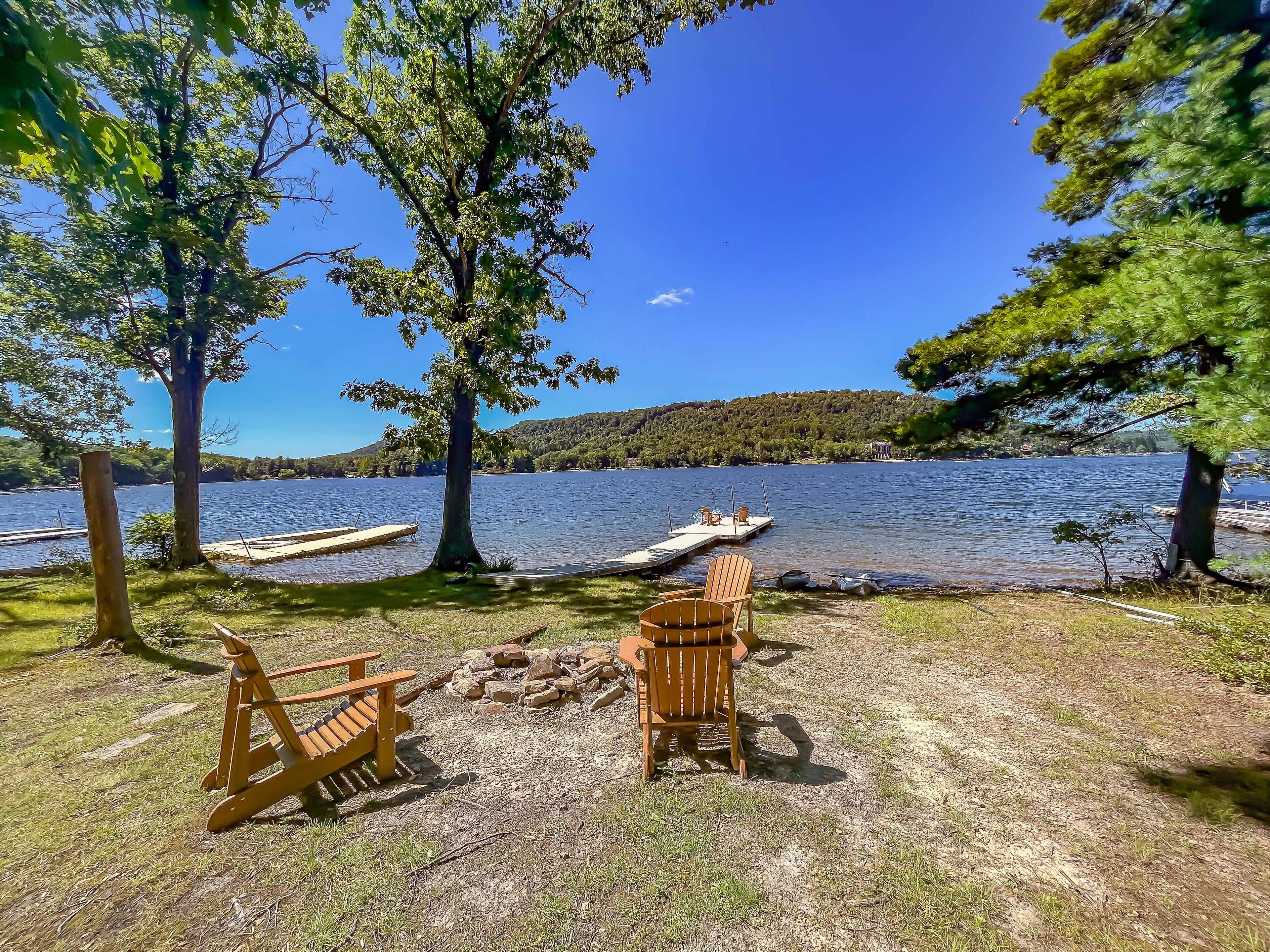 A lakeside setting with wooden chairs and a fire pit, surrounded by trees and a dock with boats.