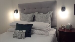Ein ordentlich bezogenes Bett mit gemusterten Kissen und einem Zierkissen, ein Nachttisch mit einer Uhr und einer Schachtel sowie Wandleuchten.