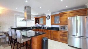 A modern kitchen with a central island, stainless steel appliances, wooden cabinets, and a black trash bin. - Stunning Beachfront Condo on Payne's Bay (Paynes Bay)
