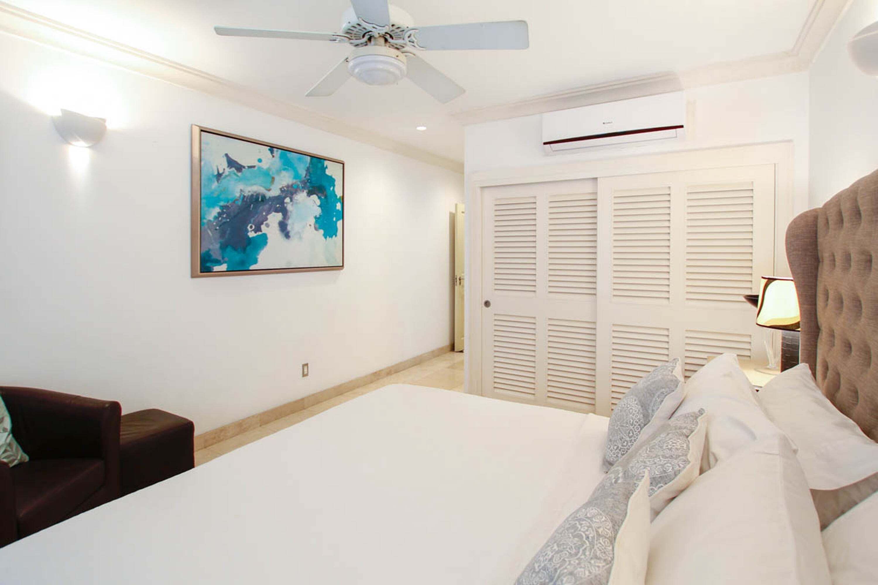 Quarto de hotel com uma cama, ventoinha no teto, ar condicionado, um quadro na parede e uma porta com persianas.
