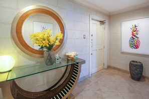 Un pasillo con una mesa consola de vidrio, un espejo redondo, un jarrón con flores amarillas, una puerta y una obra de arte colgada en la pared.